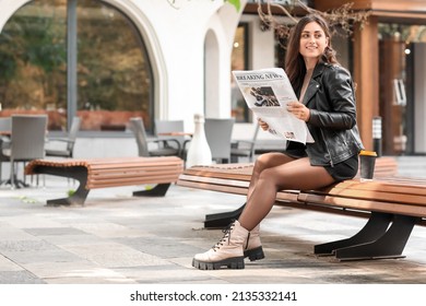 Stylish young woman reading newspaper on bench outdoors