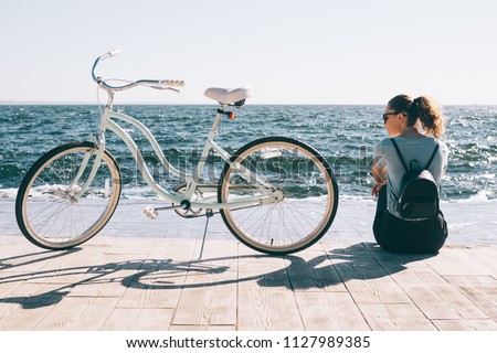 Stylish young woman sitting at the background of blue sea looking at her shiny cruiser bicycle. Female wearing backpack and casual outfit resting after bike ride at sunny summer day.