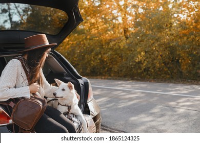 Stylish Young Woman Sitting With Cute White Dog In Car Trunk On Background Of Sunny Autumn Trees. Road Trip With Pet. Traveling With Swiss Shepherd Puppy. Space For Text