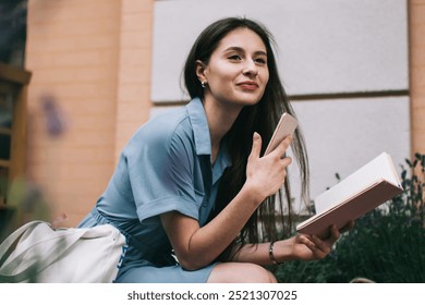 Stylish young woman with smartphone and planner looking away while sitting near baskets with flowers and studying on city street - Powered by Shutterstock