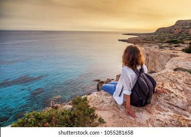 A Stylish Young Woman Traveler Watches A Beautiful Sunset On The Rocks On The Beach, Cyprus, Cape Greco, A Popular Destination For Summer Travel In Europe