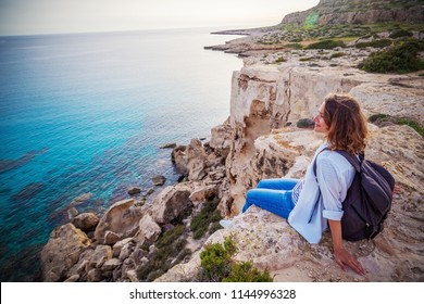 A Stylish Young Woman Traveler Watches A Beautiful Sunset On The Rocks On The Beach, Cyprus, Cape Greco, A Popular Destination For Summer Travel In Europe