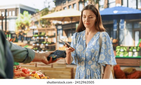 Stylish Young Woman Using Smartphone with Contactless NFC Payment Technology to Buy Organic Vegetables at a Farmers Market. Street Vendor Holding an Electronic Online Payment Terminal Device - Powered by Shutterstock