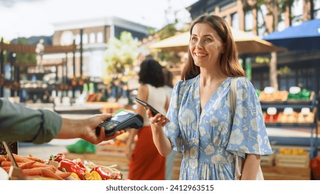 Stylish Young Woman Using Smartphone with Contactless NFC Payment Technology to Buy Organic Vegetables at a Farmers Market. Street Vendor Holding an Electronic Online Payment Terminal Device - Powered by Shutterstock