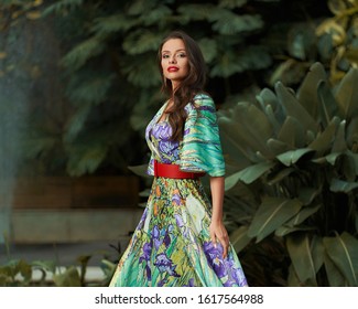 Stylish young woman wearing luxury colourful dress posing at summer garden looking at camera. Female fashion model relaxing outdoor at green tropical plant background motion shot