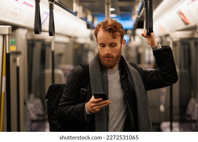 Stylishly dressed bearded man stands in a subway car in headphones and a smartphone in his hand. Passenger in public transport - Powered by Shutterstock