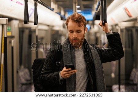 Stylishly dressed bearded man stands in a subway car in headphones and a smartphone in his hand. Passenger in public transport