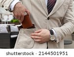A stylishly dressed business man puts a brown leather wallet in his jacket pocket. Horizontal orientation, no face 
