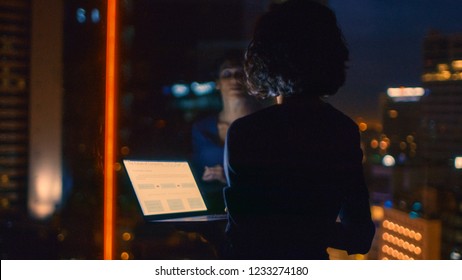 Stylishly Dressed Businesswoman Holds Laptop While Looking Out Of The Window Of Her Office. Late At Night Woman Doing Important Job. Window Has Big City Business District View With Many Night Lights.