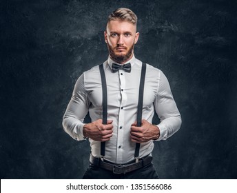 Stylishly Dressed Young Man In Shirt With Bow Tie And Suspenders. Studio Photo Against A Dark Wall Background