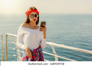 Stylishly dressed young woman listens to music in earphones and uses smartphone on beach against sea background. Street Fashion