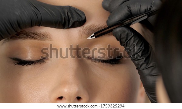 Stylist\'s hands in\
black gloves plucking eyebrows with tweezers. Beautiful attractive\
female face of a blonde well-groomed woman or lady. Styling and\
lamination of eyebrows.\
