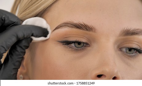 Stylist's hands in black gloves wipe the model face with a cotton pad. Beautiful attractive female face of a blonde well-groomed woman or lady. Styling and lamination of eyebrows. 