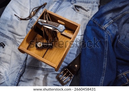 Stylized composition of a folded pocket knife, compact flashlight, and paper money placed inside a wooden box, all set on denim fabric.