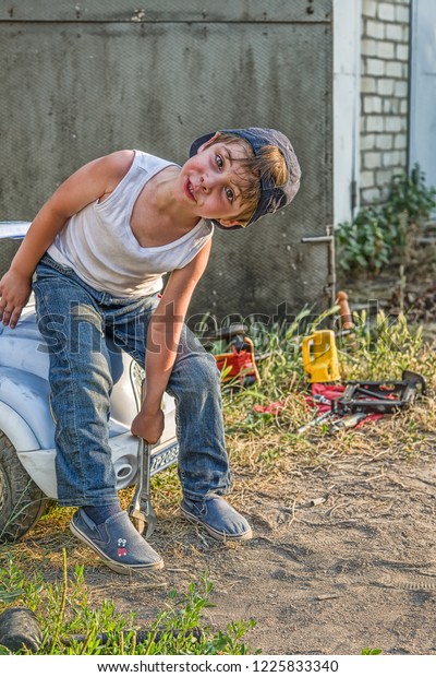 Stylized foto of a boy in a tank,\
jeans and a baseball cap in the garage repairing the\
car.