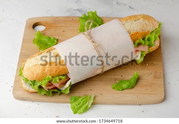 Sub deli sandwich baguette with\
ham, cheese, tomatoes and lettuce top view on cutting\
board