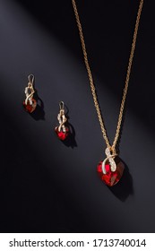 Subject shot of  jewelry set composed of two golden earrings and a necklace with ruby heart-shaped gems and white crystals. The elegant jewels are located on the black surface. 