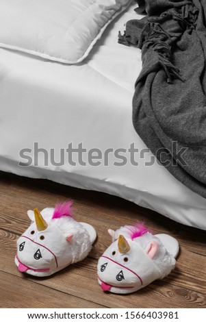 Subject shot of plush house slippers made in the form of white teasing unicorn. The slippers are next to the bed with white linen, pillow and a gray plaid on it. 