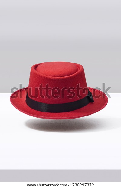 Subject shot of a red porkpie\
hat decorated with a black silk band with a bow. The stylish hat is\
hovering in the air under the white surface against the gray\
backdrop.