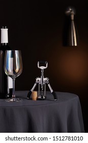 Subject Shot Of A Steel Winged Butterfly Corkscrew With Plastic Handles And A Cork Cap Near It, A Dark Glass Bottle And A Wine Glass With A Mirroring Effect. 