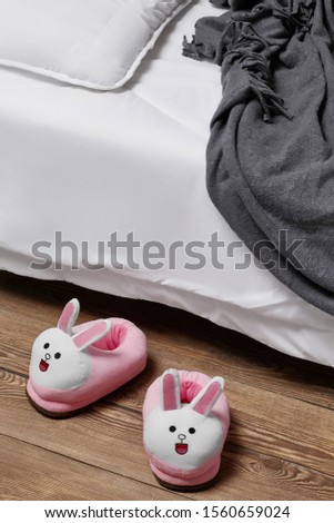 Subject shot of white and pink plush house slippers made in the form of kawaii rabbit. The slippers are next to the bed with white linen, pillow and a gray plaid on it. 