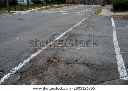 Suburban asphalt road intersection featuring a bicycle lane symbol, white and yellow dividing lines, a metal manhole cover, and scattered dry autumn leaves on cracked pavement within a residential neighborhood, depicting urban infrastructure.