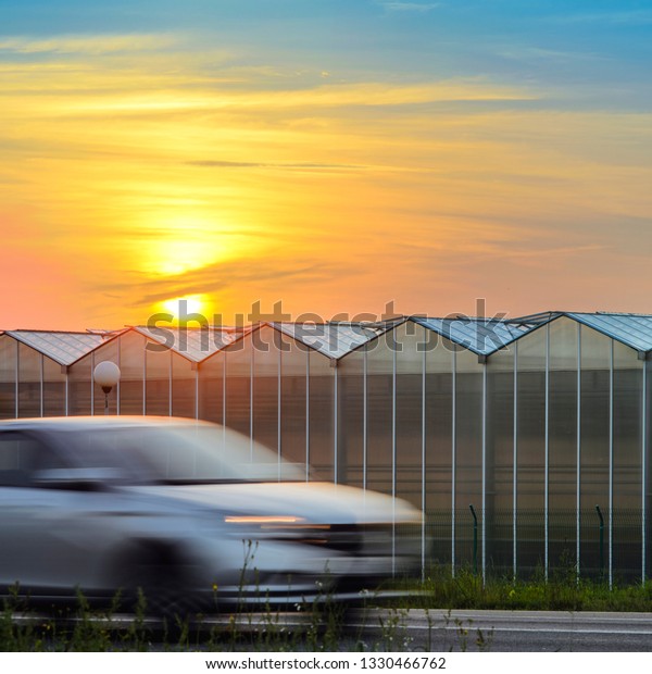 Suburban Highway Near the Greenhouse Plant at\
Sunset. Driving Cars Blurred in Motion. Large Industrial Greenhouse\
at Sunset. Gorgeous Sunset Red and Orange Sky Over the Building of\
Greenhouses Plant.