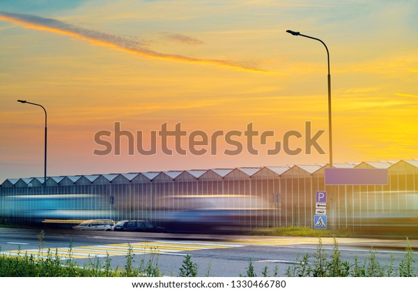 Suburban Highway Near the Greenhouse Plant at\
Sunset. Driving Cars Blurred in Motion. Large Industrial Greenhouse\
at Sunset. Gorgeous Sunset Red and Orange Sky Over the Building of\
Greenhouses Plant.