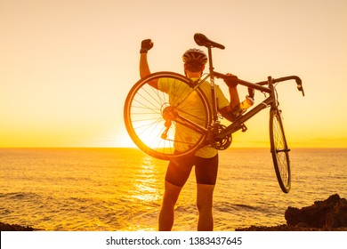 Success, achievement and winning concept with cyclist man road biking. Happy male professional athlete cycling raising arms lifting bike by sea during sunset cheering and celebrating at summit top.