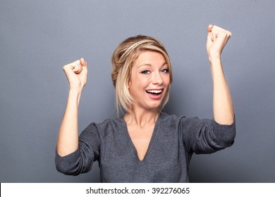 Success Concept - Happy Young Blond Woman Winning A Competition With Fun Body Language And Hands Up, Gray Background Studio