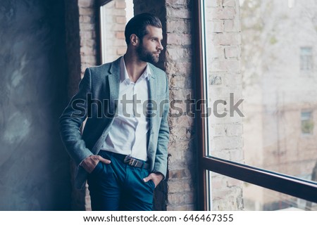 Success concept. Stylish young bearded man is standing near the window and looking far. He is in a suit and, with hands in pockets, pensive and concentrated