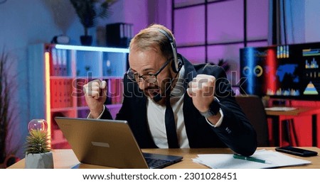 Success and luck concept where likable surprised professional bearded businessman in headset getting good news on laptop screen and rejoycing with raising hands