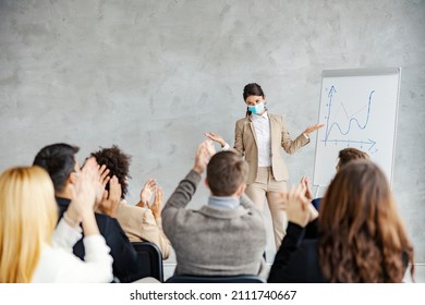 Success And Stock Market Raise During Covid. A Female Broker With A Face Mask On Her Face Standing Next To A Chart Board With Analysis And Having Presentation While Her Colleagues Clapping.