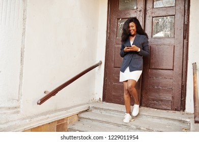 Success Stylish African American Woman In Jacket And Skirt Against Old Wooden Door With Smartphone At Hands.