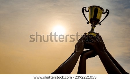 Success of teamwork, joint achievement of goal in business and life. Winning team is holding trophy in hands. Silhouettes of many hands in sunset.