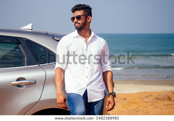 success traveler driver\
indian model male in a white shirt freelancer smiling and posing\
seacost .Handsome bearded man is standing near car freelancing on\
the beach