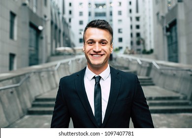 Success In Work. Beautiful Young Cheerful Businessman In A Smart Black Suit And Tie With A Big Genuine Smile Is Looking Directly At The Camera.