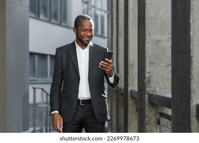 A successful African American businessman in a business suit is standing on the street near a skyscraper wearing a suit and using a mobile phone.