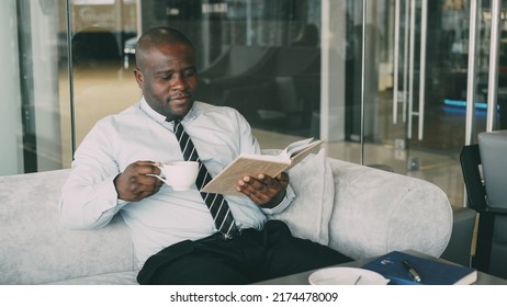 Successful African American businessman in formal clothes reading book while drinking coffee in glassy cafe at lunch break - Powered by Shutterstock