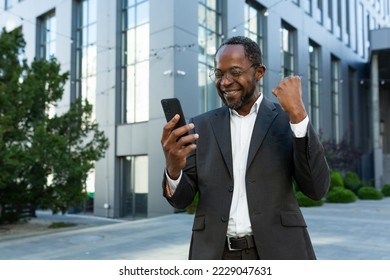 Successful african american mature investor with phone outside office building reading happy news online, senior businessman celebrating victory in business suit and glasses. - Powered by Shutterstock