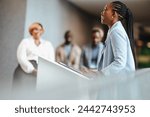 Successful African business woman engages with her audience while giving an inspiring speech at a professional conference. Colleagues listen intently in a modern corporate setting.