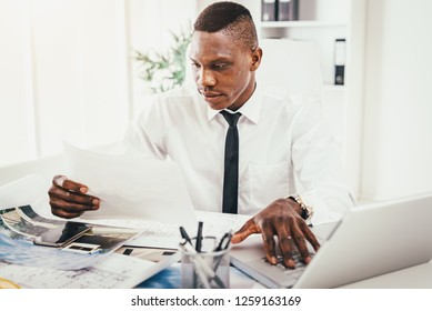 Successful  African businessman looking on paper document and working on laptop in modern office. - Shutterstock ID 1259163169