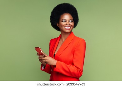 Successful african businesswoman hold smartphone look aside with confident happy smile. Afro american young entrepreneur female in luxury red suit with mobile phone isolated over green studio wall - Shutterstock ID 2033549963