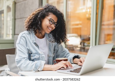 Successful african-american young woman girl student freelancer working remotely online on laptop, doing homework, project on distance outdoors in cafe