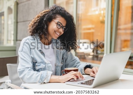 Successful african-american young woman girl student freelancer working remotely online on laptop, doing homework, project on distance outdoors in cafe