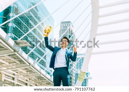 Successful Asian business man hand holding golden trohpy up in the city office building background, Smart working, Success people, Partner and Teamwork concept
