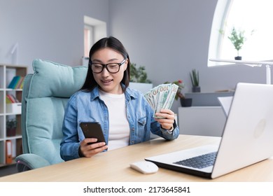 Successful Asian Business Woman Is Happy With Winning In Online Casino, Looking At Phone Screen And Holding Money Dollars In Hands, Business Woman Working In Office.