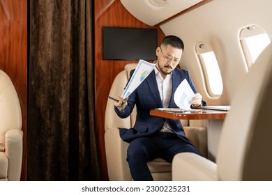 successful Asian businessman in suit and glasses sits in private jet and works with documents, Korean financier manager writes report and charts in airplane, person works in business class - Powered by Shutterstock