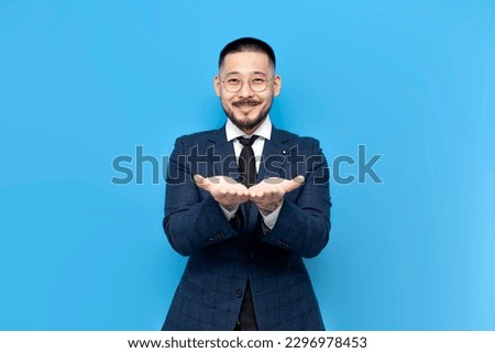 successful asian businessman in suit holds his hands in front of him on blue isolated background, korean entrepreneur holds nothing in his palms and gives empty space