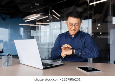 Successful asian businessman use smart watch at workplace, man chooses apps and smile happy with apps, employee inside office use laptop at work. - Powered by Shutterstock